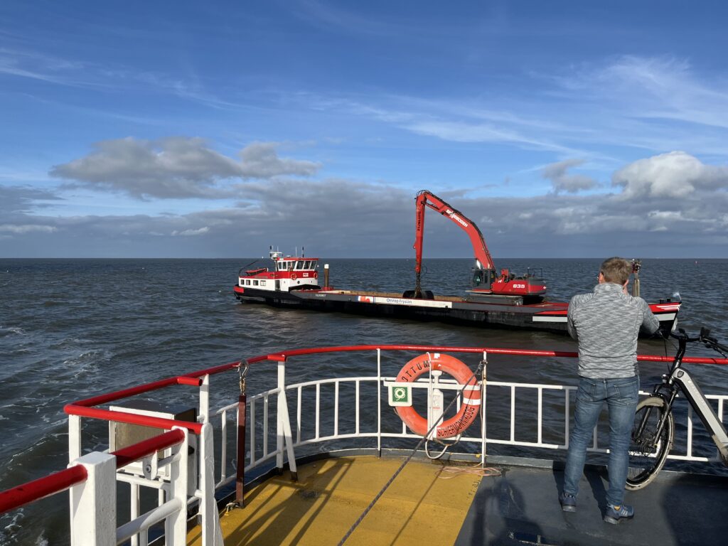 Zicht vanaf het dek van een veerboot. Naast de boot vaart een schip met een graafmachine aan boord